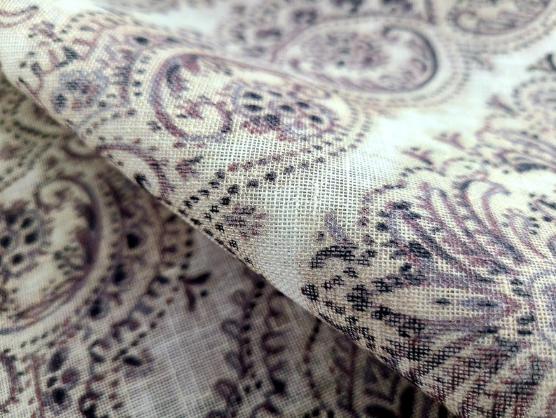 May include: Close-up of a beige fabric with a paisley pattern in shades of brown and black. The fabric has a textured, woven look.