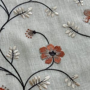 K&ouml;nnte beinhalten: Nahaufnahme eines beigefarbenen Stoffes mit gestickten Blumenmustern in Braun-, Orange- und Schwarzt&ouml;nen. Die Blumen haben ein schlichtes, zartes Design mit f&uuml;nf Bl&uuml;tenbl&auml;ttern.