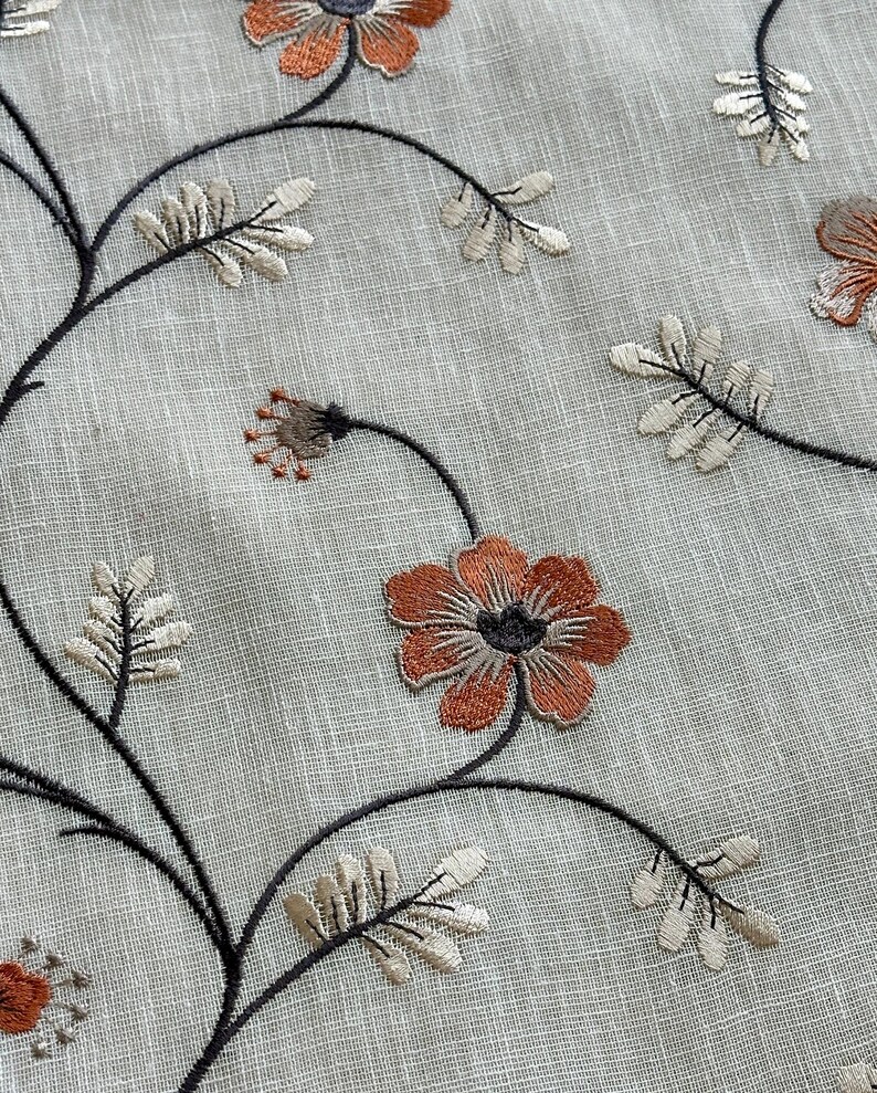 K&ouml;nnte beinhalten: Nahaufnahme eines beigefarbenen Stoffes mit gestickten Blumenmustern in Braun-, Orange- und Schwarzt&ouml;nen. Die Blumen haben ein schlichtes, zartes Design mit f&uuml;nf Bl&uuml;tenbl&auml;ttern.