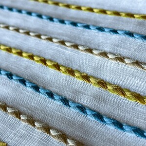 May include: White fabric with a repeating pattern of blue, yellow, and beige braided trim.