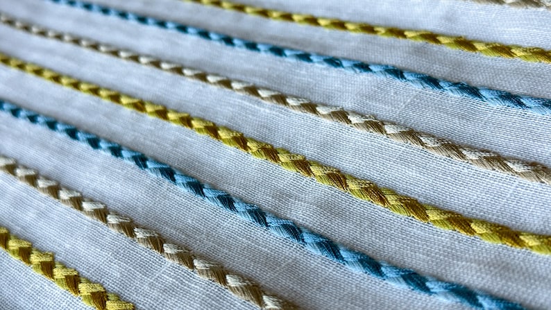 May include: White fabric with a repeating pattern of blue, yellow, and beige braided trim.