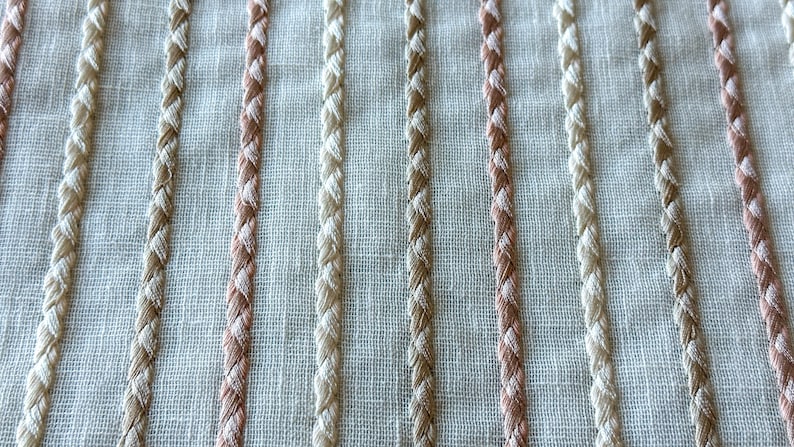 Puede incluir: Primer plano de una tela blanca con rayas verticales de hilo trenzado beige y rosa claro.