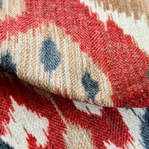 May include: Close-up of a fabric with a red, brown, and blue ikat pattern. The fabric has a textured surface and a subtle herringbone weave.