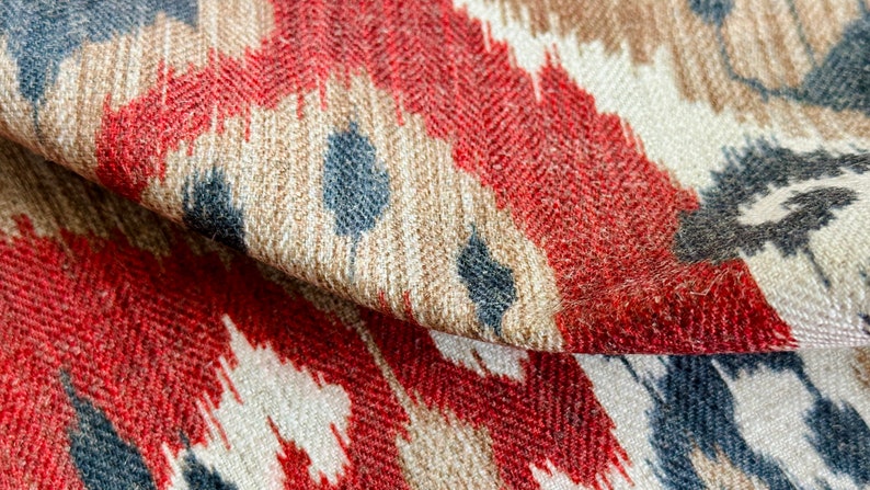 May include: Close-up of a fabric with a red, brown, and blue ikat pattern. The fabric has a textured surface and a subtle herringbone weave.