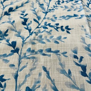 May include: Close-up of a fabric with a repeating pattern of blue watercolor-style branches and leaves against a neutral beige background. The fabric has a linen-like texture, creating a natural and organic feel. The design is suitable for home decor.