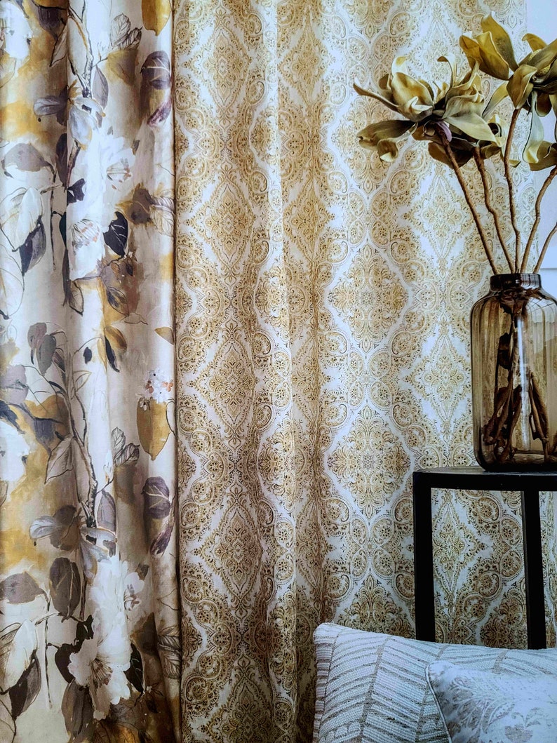 May include: Two patterned curtains, one with a floral design and the other with a paisley design, hang in a window. A vase with dried branches sits on a black table in front of the curtains.