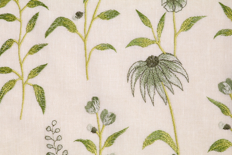 May include: A white fabric with an embroidered floral pattern. The flowers are green and white with a light brown center. The fabric has a subtle texture.