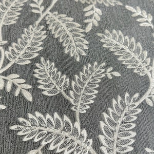 K&ouml;nnte beinhalten: Nahaufnahme eines grauen Stoffes mit einem wei&szlig;en Blumenmuster. Das Muster zeigt zarte, sich wiederholende farnartige Bl&auml;tter.