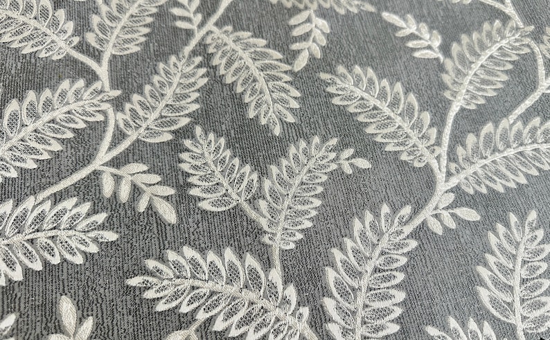 K&ouml;nnte beinhalten: Nahaufnahme eines grauen Stoffes mit einem wei&szlig;en Blumenmuster. Das Muster zeigt zarte, sich wiederholende farnartige Bl&auml;tter.