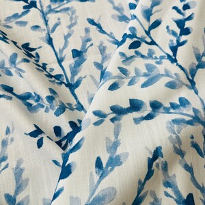 May include: Close-up of white fabric with a repeating pattern of blue watercolor-style branches and leaves. The fabric is folded, showing depth and texture. The design evokes a natural, botanical theme.