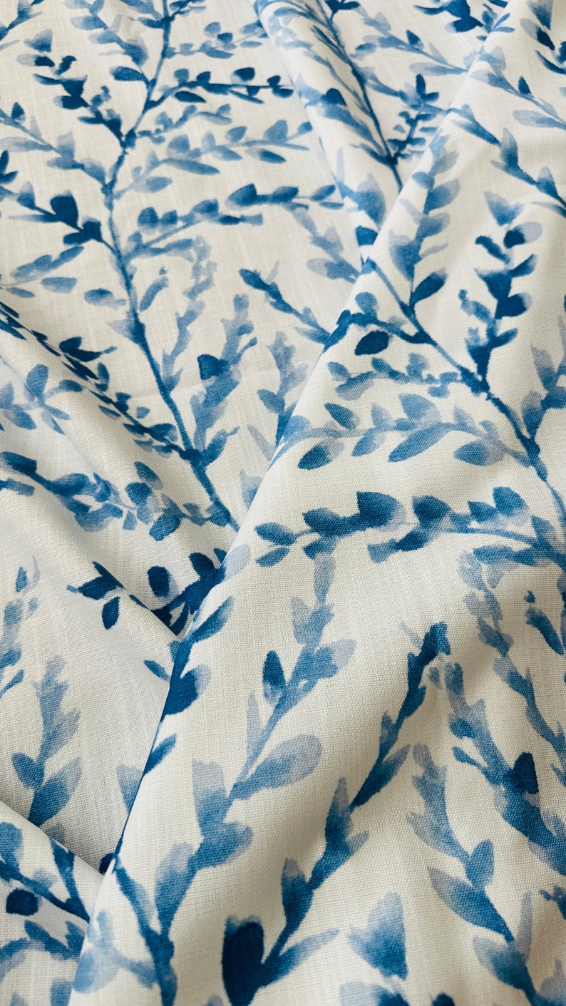 May include: Close-up of white fabric with a repeating pattern of blue watercolor-style branches and leaves. The fabric is folded, showing depth and texture. The design evokes a natural, botanical theme.