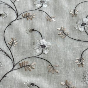 May include: A close-up of a sheer white fabric with embroidered brown floral designs. The flowers are white with brown centres and the leaves are brown.