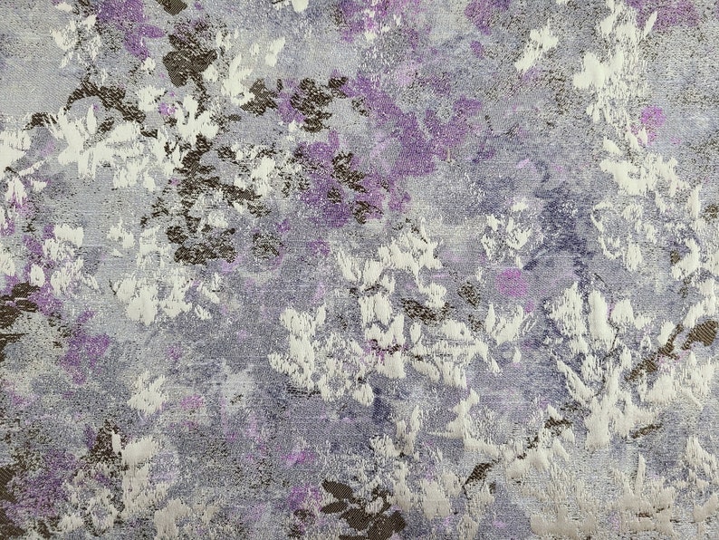 Pu&ograve; includere: Primo piano di un tessuto con un motivo floreale in tonalit&agrave; di viola, bianco e marrone. Il tessuto ha una superficie testurizzata e un leggero bagliore.