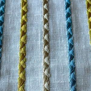 May include: Close-up of a white fabric with embroidered stripes in yellow, beige, and blue.