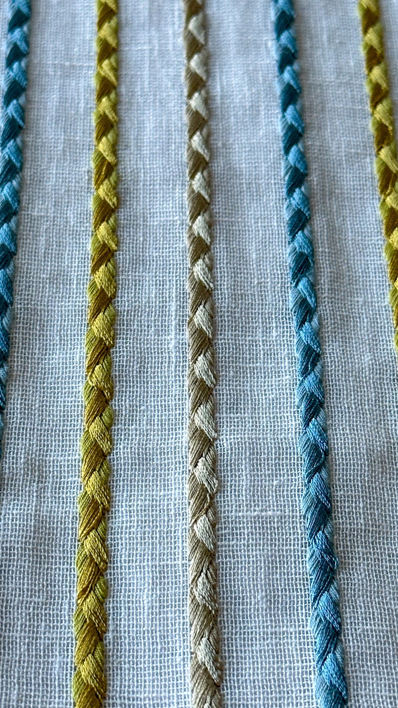 May include: Close-up of a white fabric with embroidered stripes in yellow, beige, and blue.