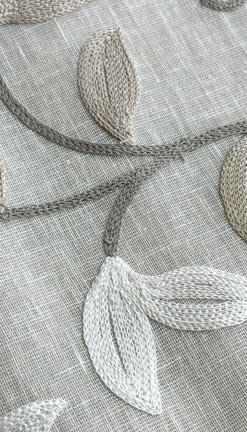 May include: Close-up of a white linen fabric with embroidered leaves in beige and white thread. The leaves are arranged in a flowing pattern.
