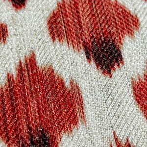 May include: Close-up of a white fabric with a red and black ikat pattern. The fabric has a textured weave and the pattern is made up of repeating shapes.