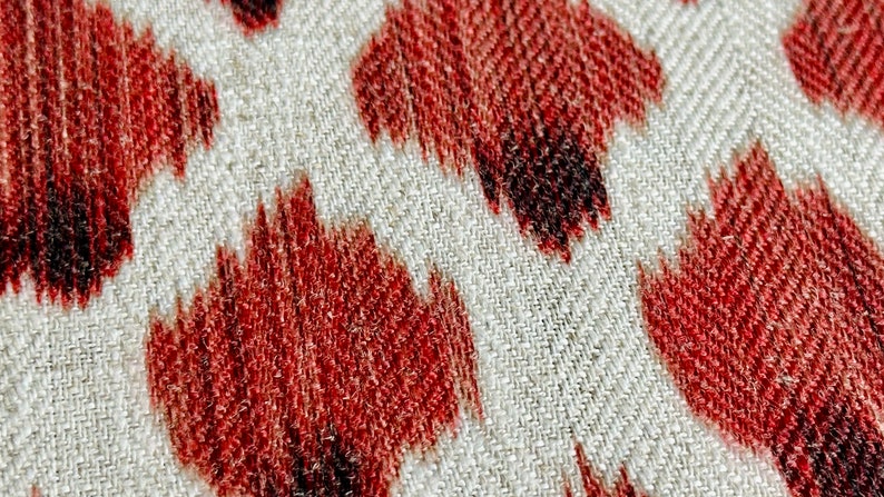 May include: Close-up of a white fabric with a red and black ikat pattern. The fabric has a textured weave and the pattern is made up of repeating shapes.