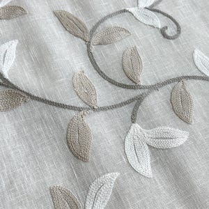 May include: Close-up of a beige fabric with embroidered leaves in shades of brown and white. The leaves are arranged in a vine-like pattern.