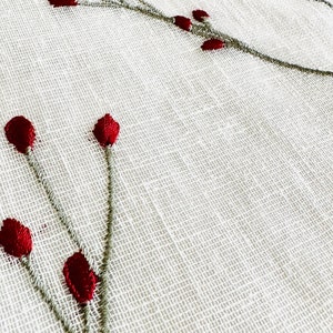 K&ouml;nnte beinhalten: Wei&szlig;er Stoff mit gestickten roten Beeren und gr&uuml;nen Stielen. Der Stoff hat eine gewebte Textur.