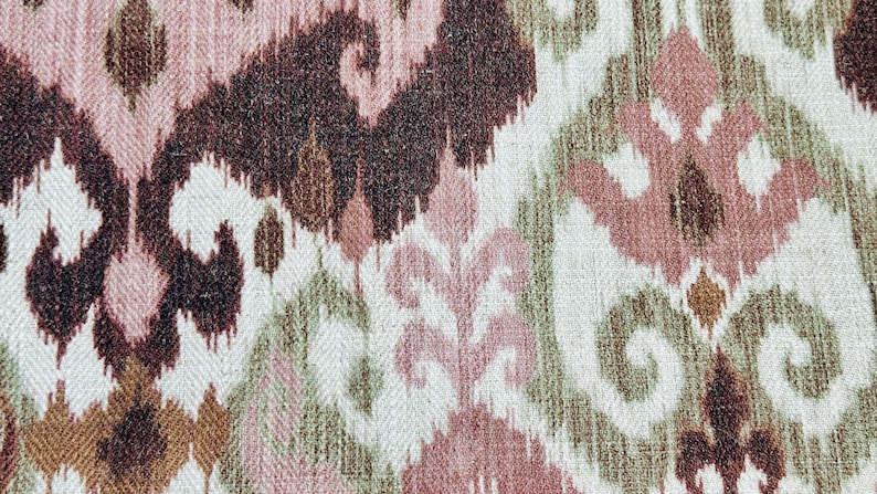May include: Close-up of a woven fabric with an ikat pattern. The design features repeating abstract shapes in shades of brown, pink, and green on a white background. The fabric has a textured appearance.
