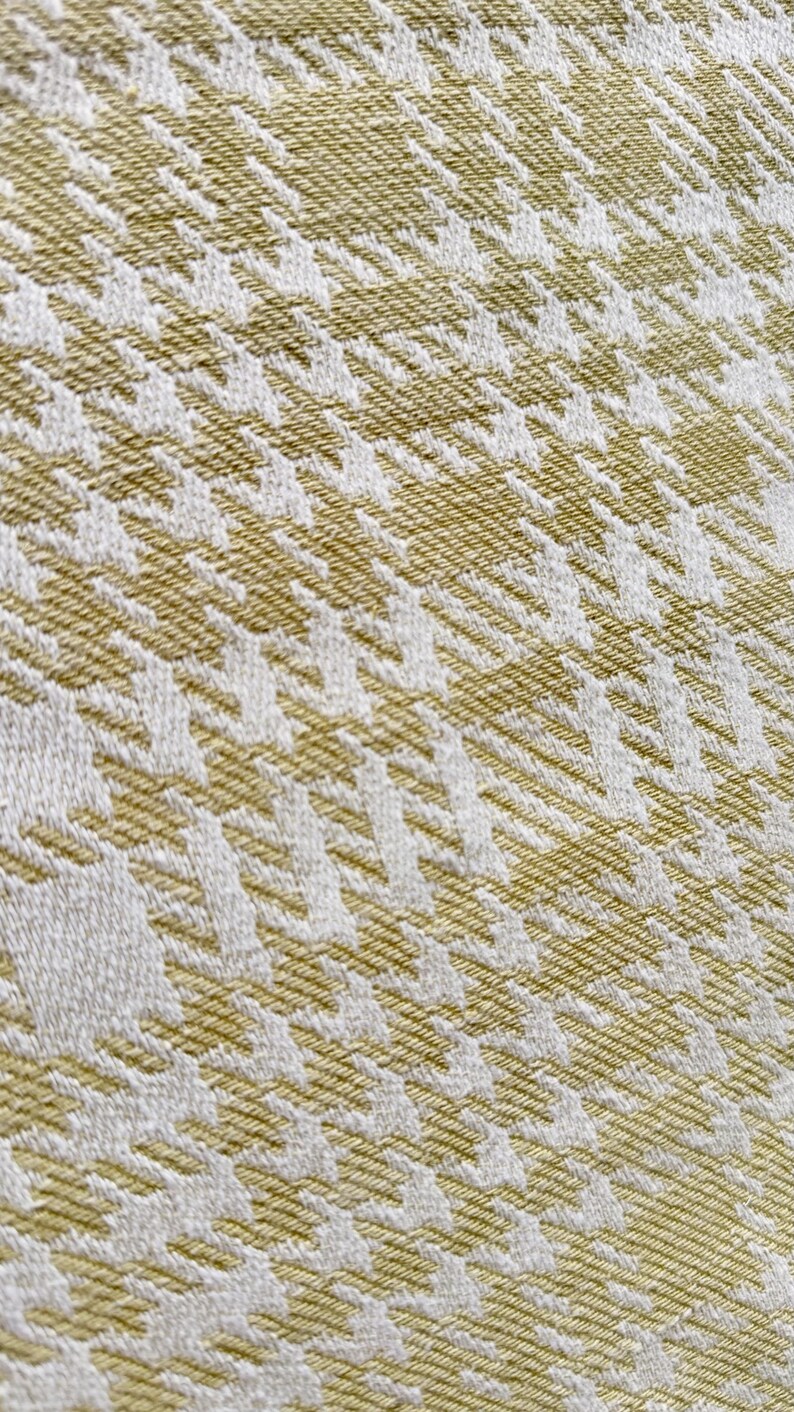 May include: A woven fabric featuring a repeating houndstooth pattern in shades of olive green and white. The textile displays a distinct diagonal texture created by the interlacing threads, showcasing a consistent geometric design throughout the surface.