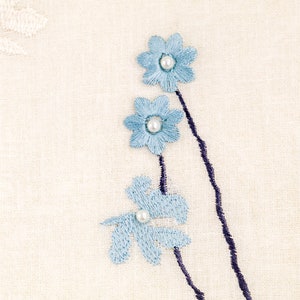 May include: White fabric with blue embroidered flowers and pearl accents. The flowers are arranged in a vertical line with a dark blue stem.