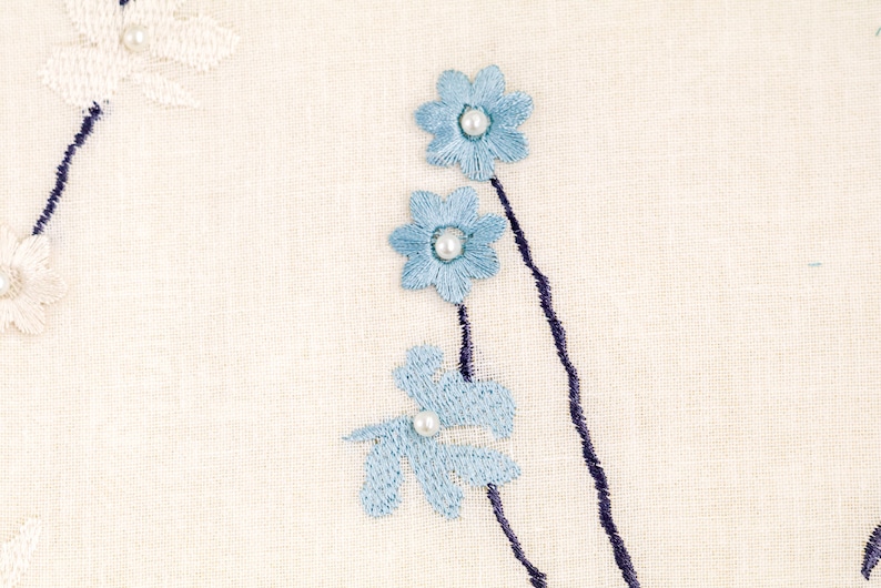 May include: White fabric with blue embroidered flowers and pearl accents. The flowers are arranged in a vertical line with a dark blue stem.