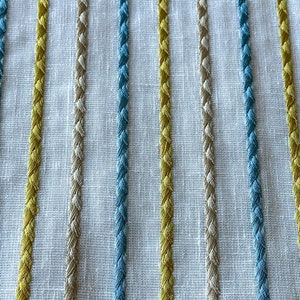 May include: Close-up of a white fabric with vertical stripes of yellow, beige, and blue thread. The stripes are woven into the fabric and create a textured look.