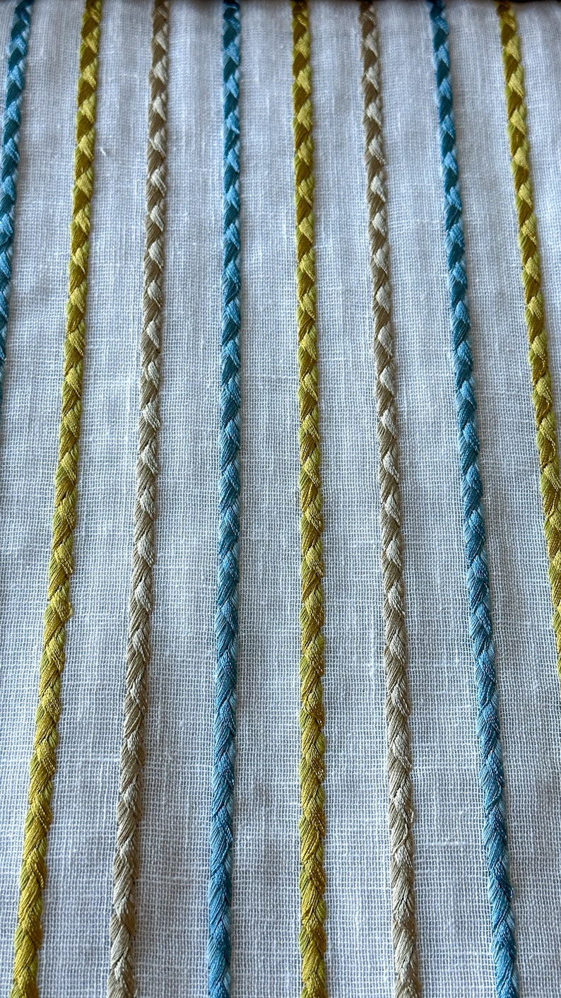 May include: Close-up of a white fabric with vertical stripes of yellow, beige, and blue thread. The stripes are woven into the fabric and create a textured look.