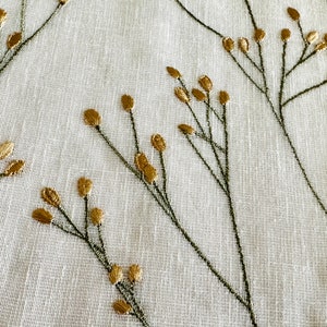 May include: White fabric with a subtle texture featuring an embroidered design of gold and green floral sprigs.
