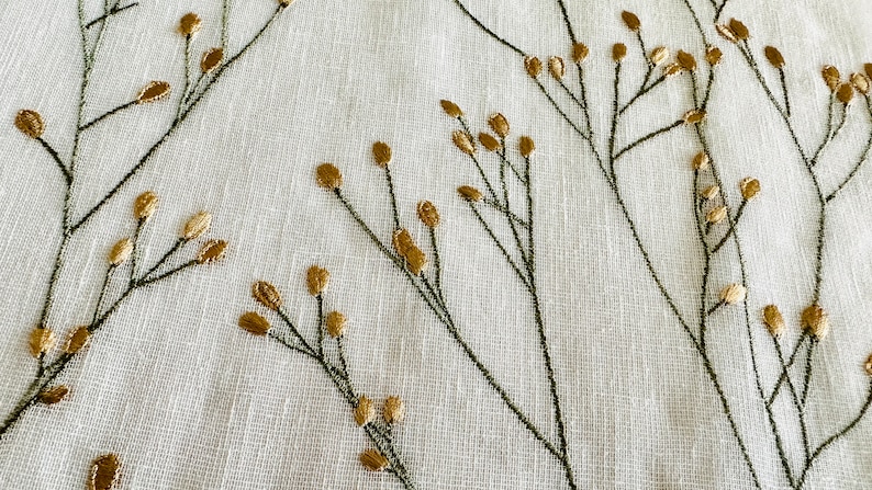 May include: White fabric with a subtle texture featuring an embroidered design of gold and green floral sprigs.
