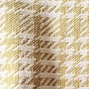 Puede incluir: Una tela tejida con un patrón repetitivo de pata de gallo en tonos amarillo dorado y blanco roto. El textil muestra una superficie texturizada con hilos entrelazados visibles, creando un diseño geométrico consistente en todo el material.