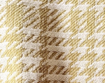 Puede incluir: Una tela tejida con un patrón repetitivo de pata de gallo en tonos amarillo dorado y blanco roto. El textil muestra una superficie texturizada con hilos entrelazados visibles, creando un diseño geométrico consistente en todo el material.