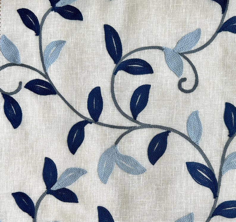 May include: A white fabric with a blue and gray embroidered floral pattern. The pattern features leaves and vines.