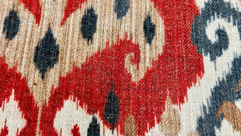May include: Close-up of a fabric with a red, white and black ikat pattern. The fabric has a textured surface and a woven appearance.