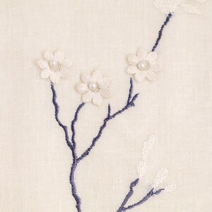 May include: A white fabric with a blue embroidered floral design. The flowers have white pearl centers.