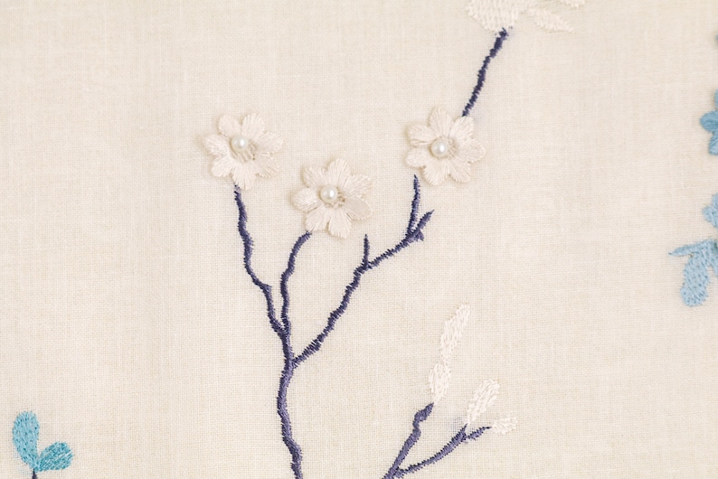 May include: A white fabric with a blue embroidered floral design. The flowers have white pearl centers.