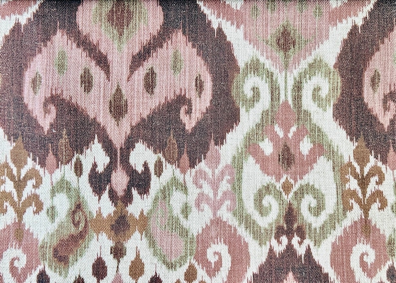 May include: Close-up of a fabric with a repeating pattern in shades of brown, pink, green, and white. The design features stylized, organic shapes reminiscent of ikat textiles, creating a textured, decorative surface.