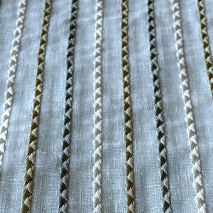 May include: White fabric with vertical stripes of different colored embroidery. The stripes are made of a braided stitch in white, gray, and brown.