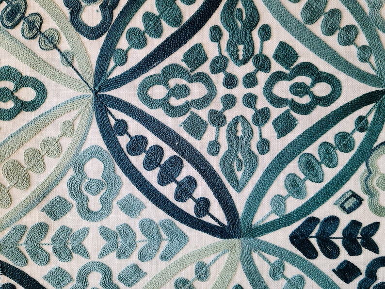 May include: Close-up of a white fabric with an intricate embroidered design in shades of blue and green. The pattern features repeating geometric shapes and floral motifs.