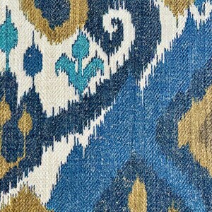 May include: A close-up of a fabric with a blue, white, and gold ikat pattern. The fabric has a woven texture and a repeating pattern of geometric shapes.