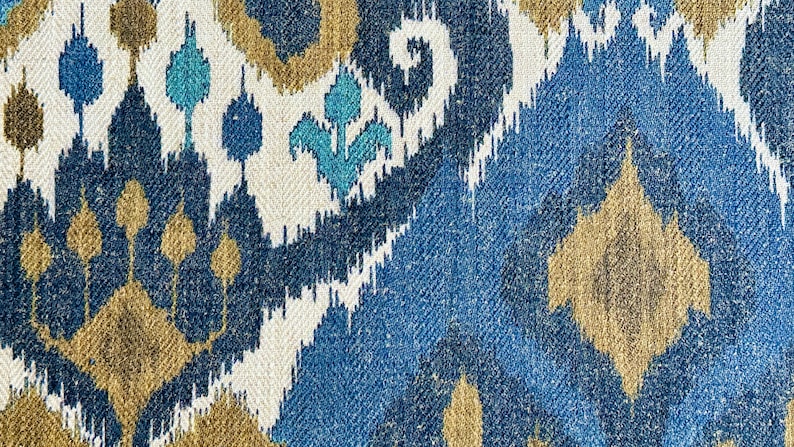 May include: A close-up of a fabric with a blue, white, and gold ikat pattern. The fabric has a woven texture and a repeating pattern of geometric shapes.
