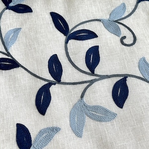May include: A close-up of a white fabric with a blue and gray embroidered floral pattern. The fabric features a repeating design of leaves and vines.