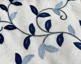 May include: A close-up of a white fabric with a blue and gray embroidered floral pattern. The fabric features a repeating design of leaves and vines.
