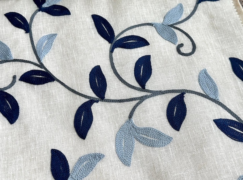 May include: A close-up of a white fabric with a blue and gray embroidered floral pattern. The fabric features a repeating design of leaves and vines.