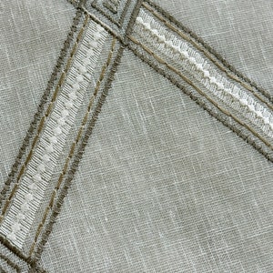 May include: A close-up of a white fabric with a brown and white embroidered design. The fabric has a subtle texture and a woven appearance.