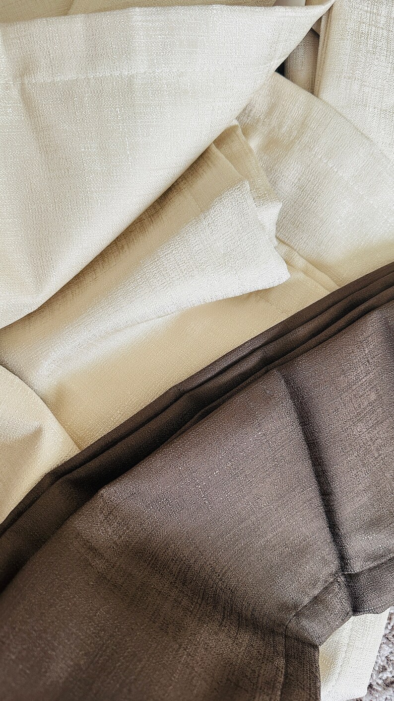 May include: Close-up of folded fabric in shades of cream and brown. The fabric has a textured appearance, with visible woven details. The folds create shadows and highlights, adding depth to the image. The overall aesthetic is simple and elegant.