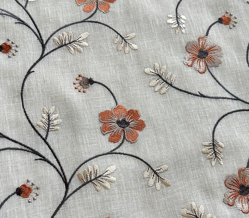 K&ouml;nnte beinhalten: Nahaufnahme eines beigefarbenen Stoffes mit gestickten Blumenmustern in Braun- und Oranget&ouml;nen. Der Stoff zeigt ein sich wiederholendes Muster aus zarten Blumen und Bl&auml;ttern.