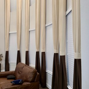 May include: Tall, elegant window curtains in a living room setting. The curtains are a combination of beige and dark brown, creating a two-tone effect. The beige fabric is at the top, transitioning to a dark brown at the bottom.